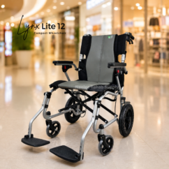 Lynx Lite 12in Wheelchair (9) A compact silver and grey lightweight Lynx lite 12 inch wheelchair is displayed prominently in the center of a bright modern shopping mall interior with softly blurred storefronts and warm ambient lighting in the background, emphasizing mobility and accessibility.